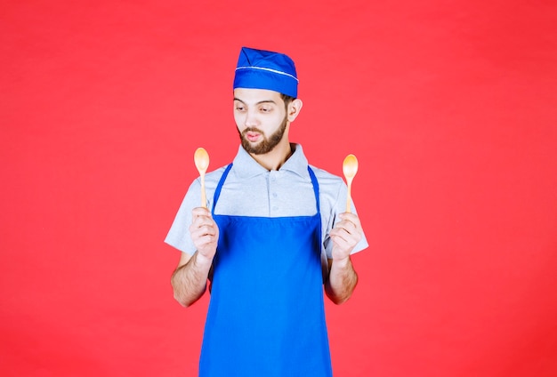 Chef in blue apron holding wooden small spoons and thinking about choice. 