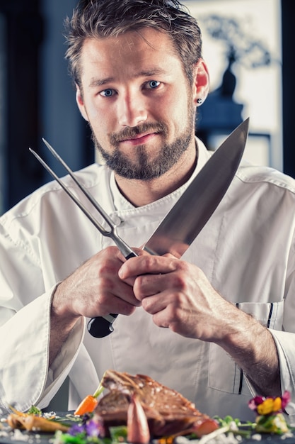 Foto chef. chef-kok grappig. chef-kok met mes en vork gekruiste armen. professionele chef-kok in een restaurant of hotel bereidt of snijdt t-bone steak. chef-kok die biefstuk bereidt. koken voor hun werk aan catering