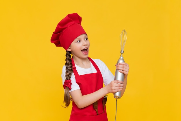 シェフの子供は泡立て用のブレンダーを持っています 赤い帽子とエプロンを着た幸せな女の子 料理の皿 黄色の孤立した背景