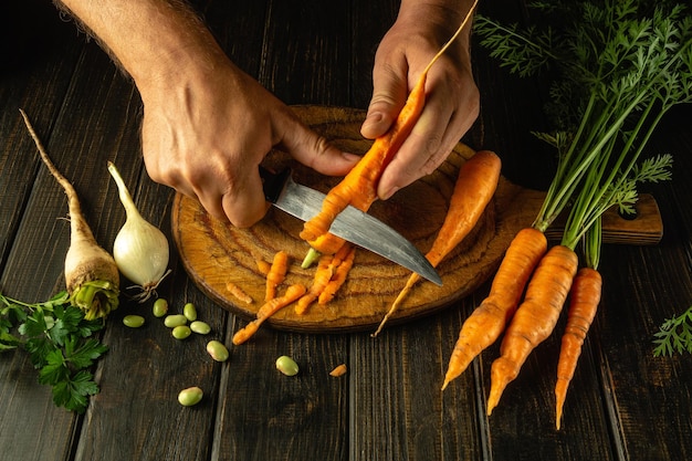 The chef cleans fresh carrots on the kitchen table before adding them to the vegetable borscht Closeup of a cook hands with a knife