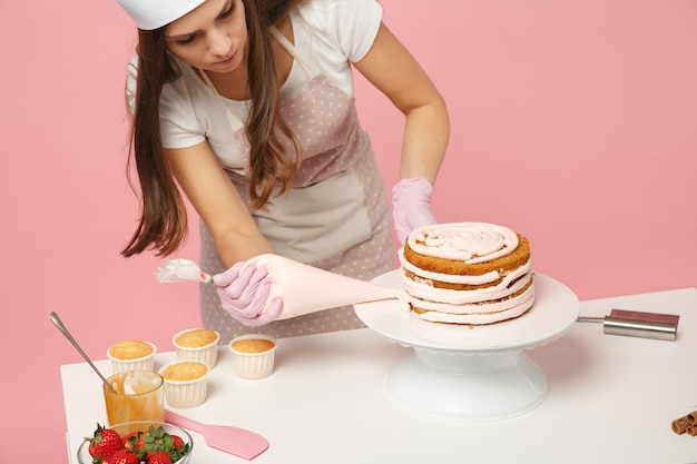 シェフは、スタジオでピンクのパステルカラーの背景に分離されたテーブルで調理する白いTシャツのトーク帽のシェフの帽子で菓子職人またはパン屋を調理します。クリーム塗布、ケーキ製造工程。コピースペースフードのコンセプトをモックアップします。