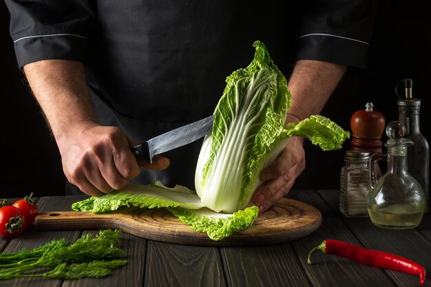 Chef or cook cuts fresh napa cabbage with knife for salad on a vintage kitchen table with vegetables