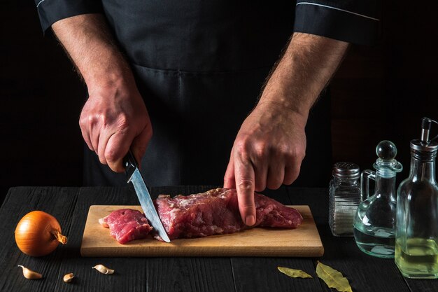 シェフまたは料理人がキッチンでナイフで牛肉を切り、料理をします。おいしいランチを作るためのキッチンテーブルの野菜とスパイス