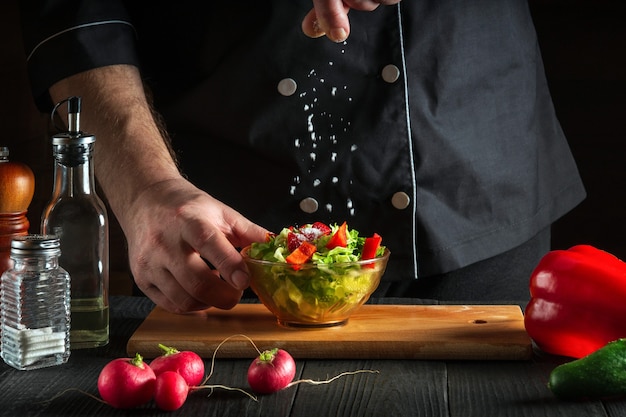 Chef or cook sprinkles salt salad of fresh vegetables on a wooden table. Preparing healthy food on the kitchen