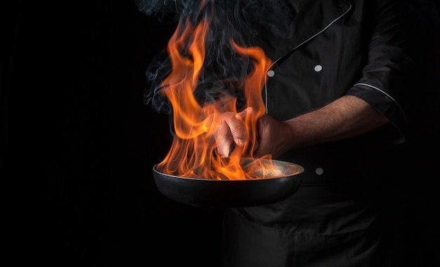 Chef cooking food in pan with fire flame on black background. Restaurant and hotel service concept. Free advertising space