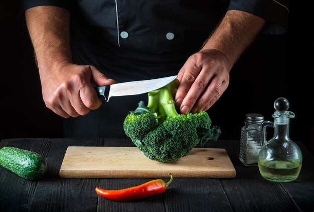 Chef cuts fresh Broccoli with a knife for salad on vintage kitchen table with fresh vegetables