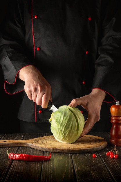 Chef cuts fresh cabbage with knife for salad on a kitchen table\
with fresh vegetables