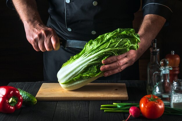 Chef cuts fresh napa cabbage with a knife for salad on a vintage kitchen table with fresh vegetables. Cooking and restaurant or cafe concept