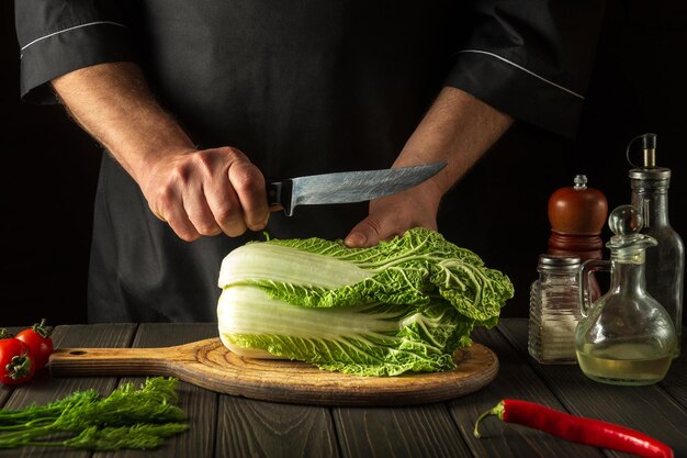 Chef cuts fresh napa cabbage with knife for salad on a vintage kitchen table with fresh vegetables