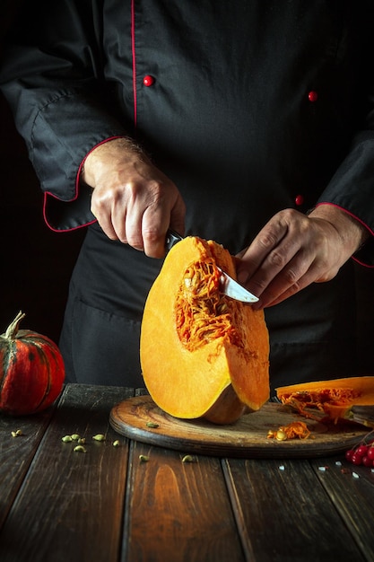 A chef cuts an orange pumpkin into slices with a knife on a wooden cutting board