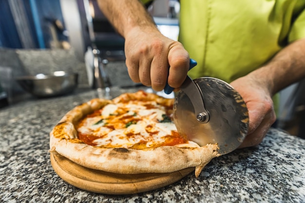 Foto chef che taglia la pizza margherita con il tagliatore di pizza su una tavola di legno in cucina