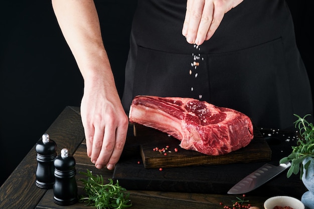 シェフ カット ステーキ ビーフ マンの手は、黒の背景に素朴な木製のまな板に生のステーキ トマホークを保持します 料理のレシピと食事の概念 選択と集中