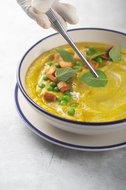 A chef in gloves decorates a soup of mashed organic vegetables with crackers, green peas and herbs with basil leaves