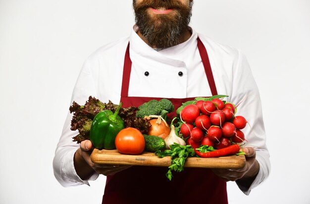 Chef holds board with fresh vegetables Cook in burgundy apron and white uniform holds salad ingredients Healthy nutrition and cuisine concept Man with beard isolated on white background