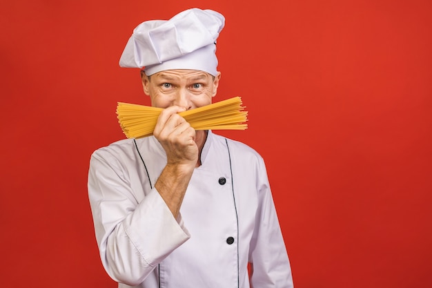 シェフは手にスパゲッティの束を保持しています。赤の背景に分離されたケータリングとイタリア料理のコンセプト。白い制服を着て満足そうな顔で料理人は乾いたパスタを握ります。