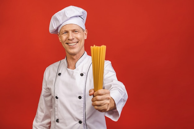 Chef holds bunch of spaghetti in hand. Catering and italian food concept isolated on red background. Cook with satisfied face in white uniform holds dry pasta.