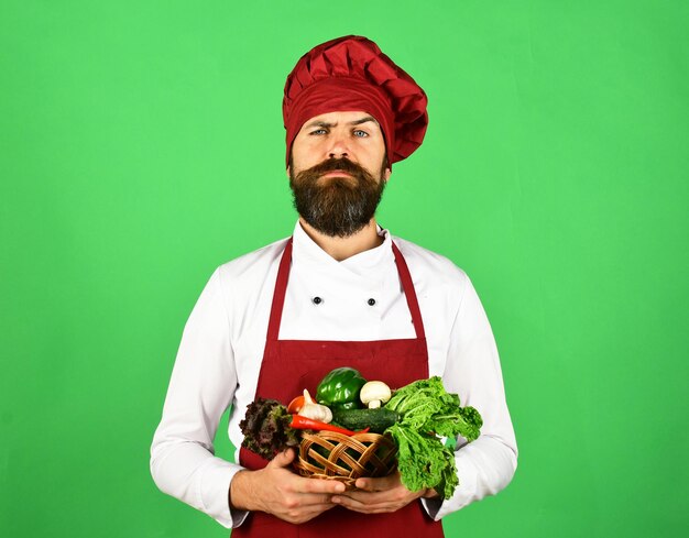 Chef holds lettuce tomato pepper and mushrooms Vegetarian restaurant concept Cook with proud face in burgundy uniform holds vegetables in wicker bowl Man with beard on green background