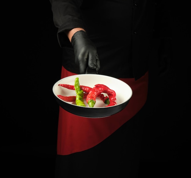 Foto chef in zwart uniform en rode schort houdt een ronde pan vast met verse chili pepers