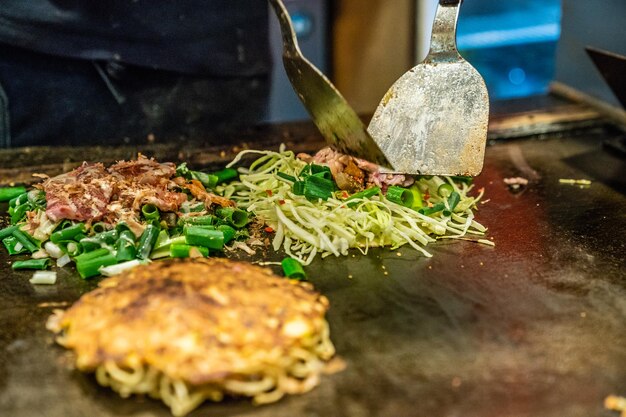요리사는 철판에 오코노미야키를 요리하기 위해 주걱을 사용하고 있으며, 그는 음식에 고기와 야채 조각을 넣습니다