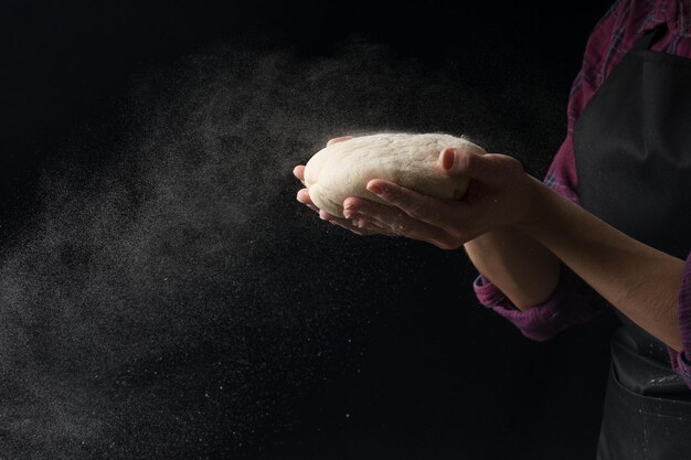 Photo the chef keeps the dough in his hands and blows the flour on a black background with an empty space under the inscription or advertisement the concept of nutrition