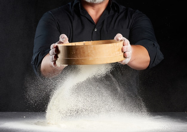 Chef-kok een man in een zwart uniform houdt een ronde houten zeef in zijn handen en zeeft wit tarwemeel