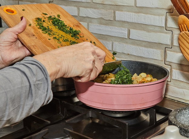 Chef-kok gooit peterselie naar courgette met wortelen zijn gebakken in koekenpan op gasfornuis