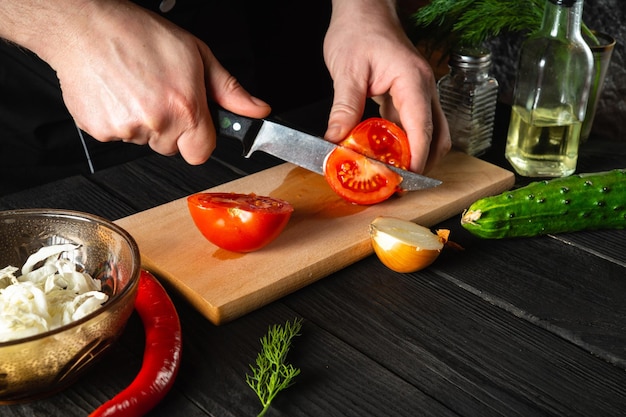 Chef-kok handen met mes, verse rode tomaten snijden voor salade. Werkomgeving in de restaurantkeuken. Verse groenten en olie op tafel