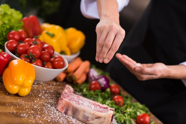 Chef-kok handen zetten zout op sappige plak rauwe biefstuk met groenten rond op een houten tafel