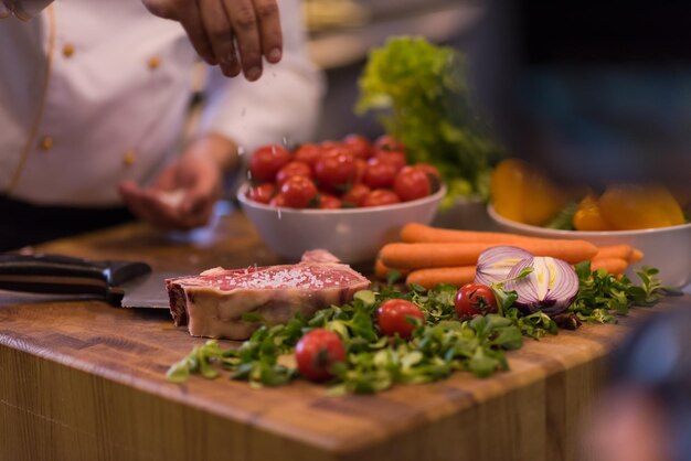 Chef-kok handen zout op sappige plak rauwe biefstuk met groenten op een houten tafel zetten