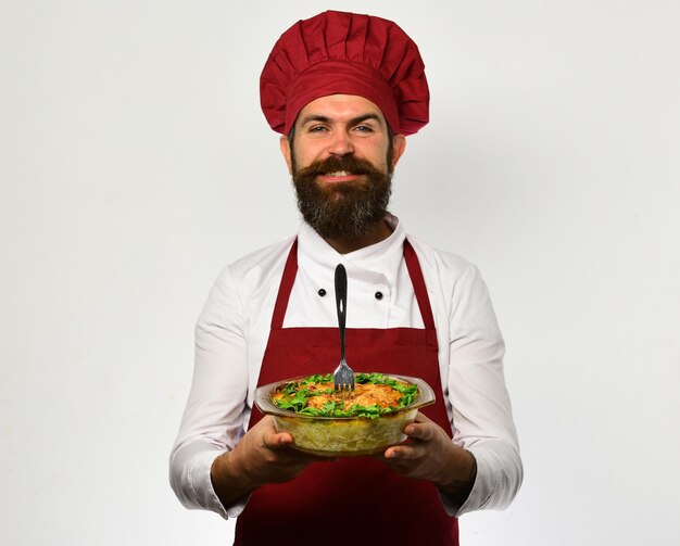 Chef-kok houdt kom met aardappelbraadpan en vork erin. Man met baard geïsoleerd op een witte achtergrond. Gezonde voeding en keuken concept. Kok met een blij gezicht in bordeauxrood uniform houdt een gebakken gerecht vast.