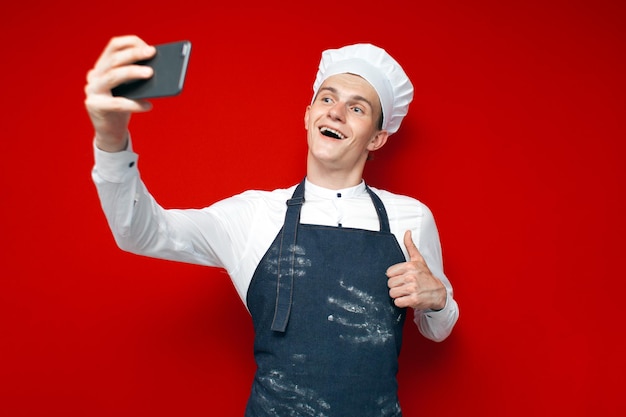 Chef-kok in uniform houdt een smartphone vast en maakt een selfie op een rode geïsoleerde achtergrond