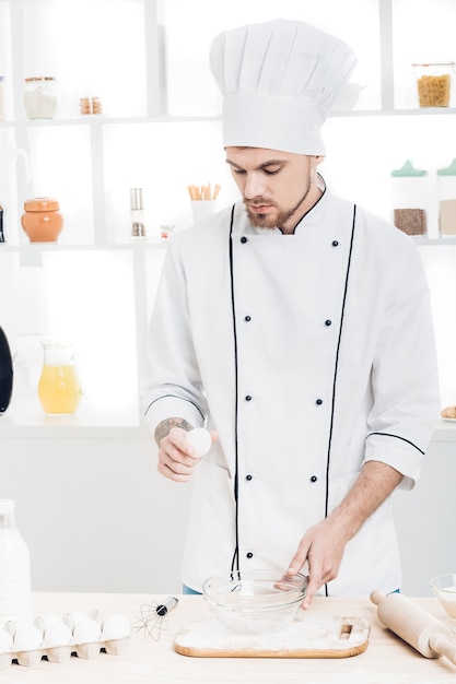 Chef-kok in uniforme breakeggs in kom om deeg in keuken te bereiden