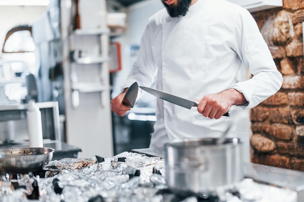 Foto chef-kok in wit uniform staat in de keuken met messen in handen