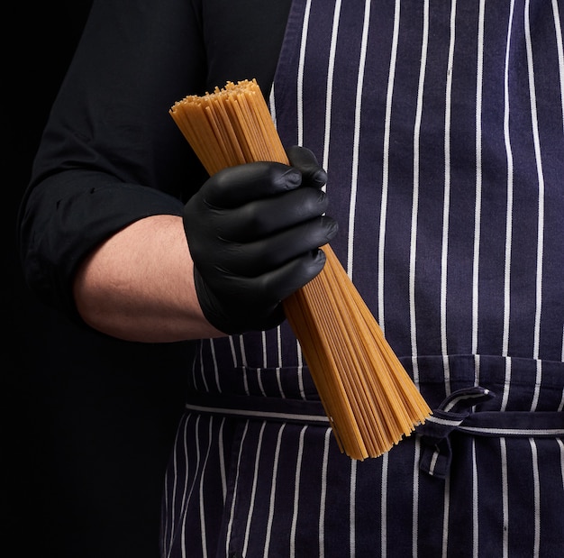 Chef-kok in zwarte latex handschoenen, gestreepte schort houdt rauwe lange gele spaghetti