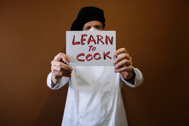 Chef-kok met een bordje met de tekst: leren en koken, hij draagt een masker op zijn gezicht