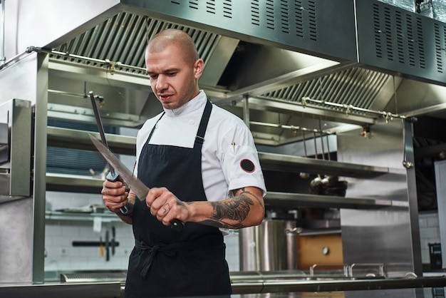 chef-kok met verschillende tatoeages op zijn armen die een mes slijpen terwijl hij in de keuken van een restaurant staat