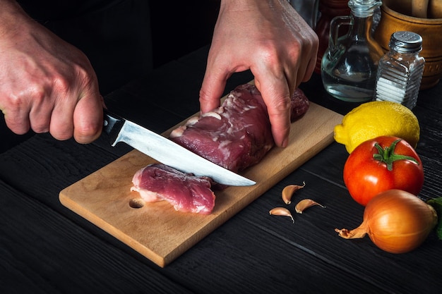 Chef-kok snijden rundvlees met mes op keuken, eten koken. Groenten en kruiden op de keukentafel om een heerlijke lunch te bereiden