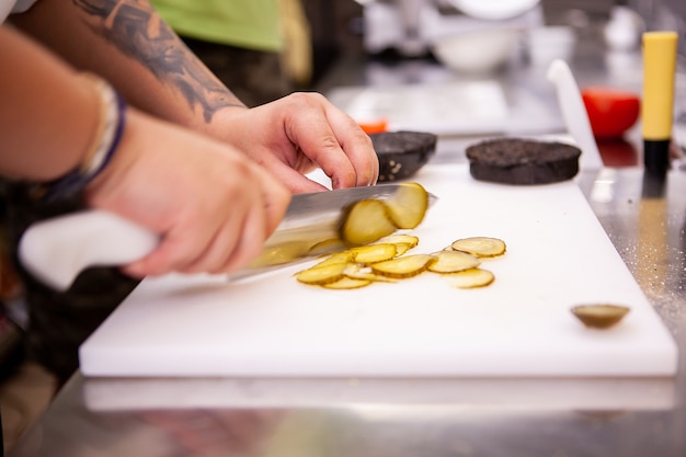 Chef-kok snijdt augurken voor heerlijke hamburger in restaurant. Voedsel voorbereiding