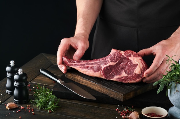 Chef-kok steak beef Mans handen houden rauwe steak Tomahawk op rustieke houten snijplank op zwarte achtergrond Koken recepten en eten concept Selectieve aandacht
