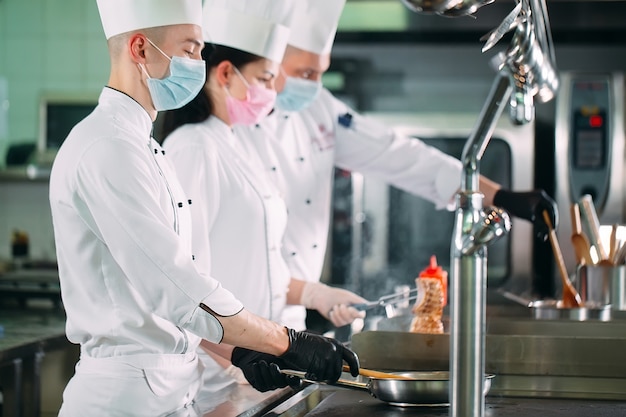 Chef-koks in beschermende maskers en handschoenen bereiden eten in de keuken van een restaurant of hotel.