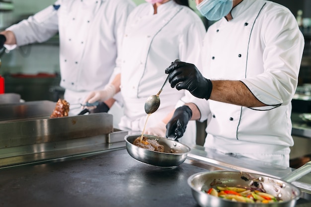 Chef-koks in beschermende maskers en handschoenen bereiden eten in de keuken van een restaurant of hotel.