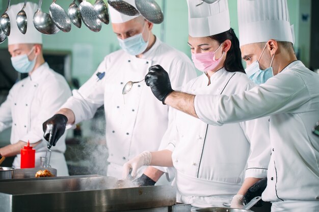 Chef-koks in beschermende maskers en handschoenen bereiden eten in de keuken van een restaurant of hotel.