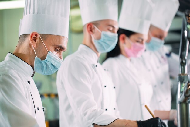 Chef-koks in beschermende maskers en handschoenen bereiden voedsel in de keuken van een restaurant of hotel.