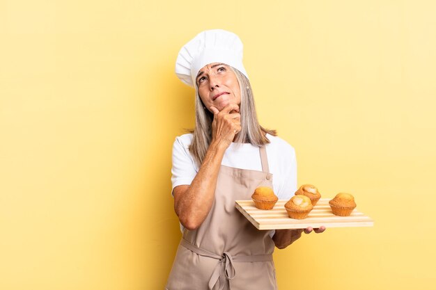 Chef-kokvrouw van middelbare leeftijd die denkt, zich twijfelachtig en verward voelt, met verschillende opties, zich afvragend welke beslissing ze moet nemen en een dienblad met muffins vasthoudt