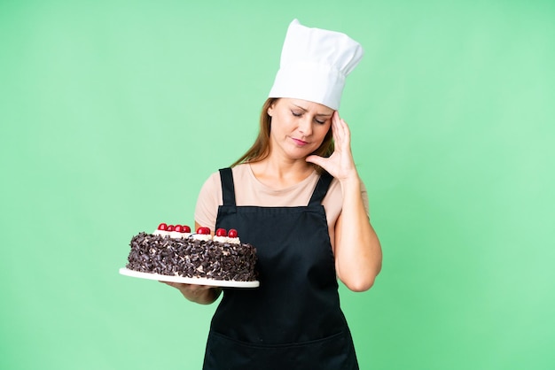 Chef-kokvrouw van middelbare leeftijd die een grote cake over geïsoleerde achtergrond met hoofdpijn houdt