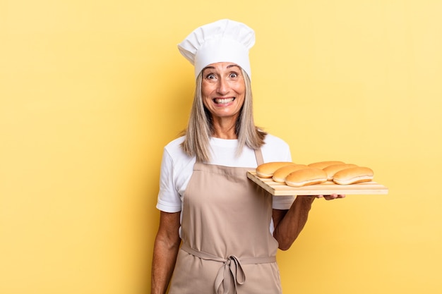 Chef-kokvrouw van middelbare leeftijd die er blij en aangenaam verrast uitziet, opgewonden met een gefascineerde en geschokte uitdrukking en een broodblad vasthoudt