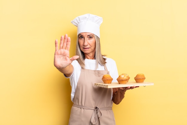 Chef-kokvrouw van middelbare leeftijd die er serieus, streng, ontevreden en boos uitziet met een open palm die een stopgebaar maakt en een dienblad met muffins vasthoudt