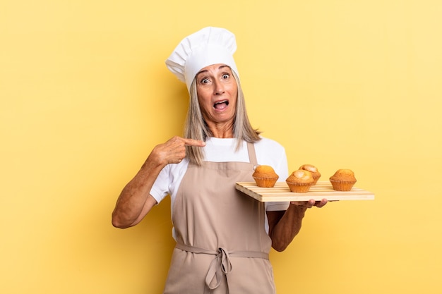 Chef-kokvrouw van middelbare leeftijd die geschokt en verrast kijkt met de mond wijd open, naar zichzelf wijst en een dienblad met muffins vasthoudt