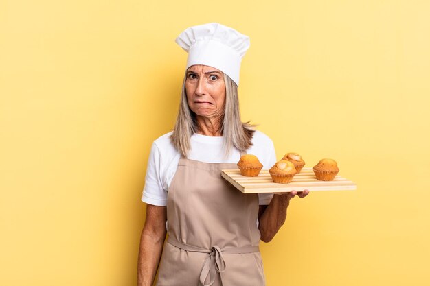 Foto chef-kokvrouw van middelbare leeftijd die verbaasd en verward kijkt, lip bijt met een nerveus gebaar, het antwoord op het probleem niet weet en een dienblad met muffins vasthoudt