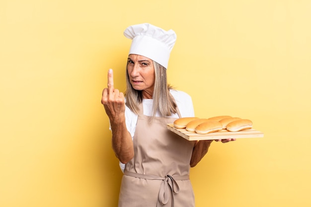 Chef-kokvrouw van middelbare leeftijd die zich boos, geïrriteerd, opstandig en agressief voelt, de middelvinger omdraait, terugvecht en een broodblad vasthoudt